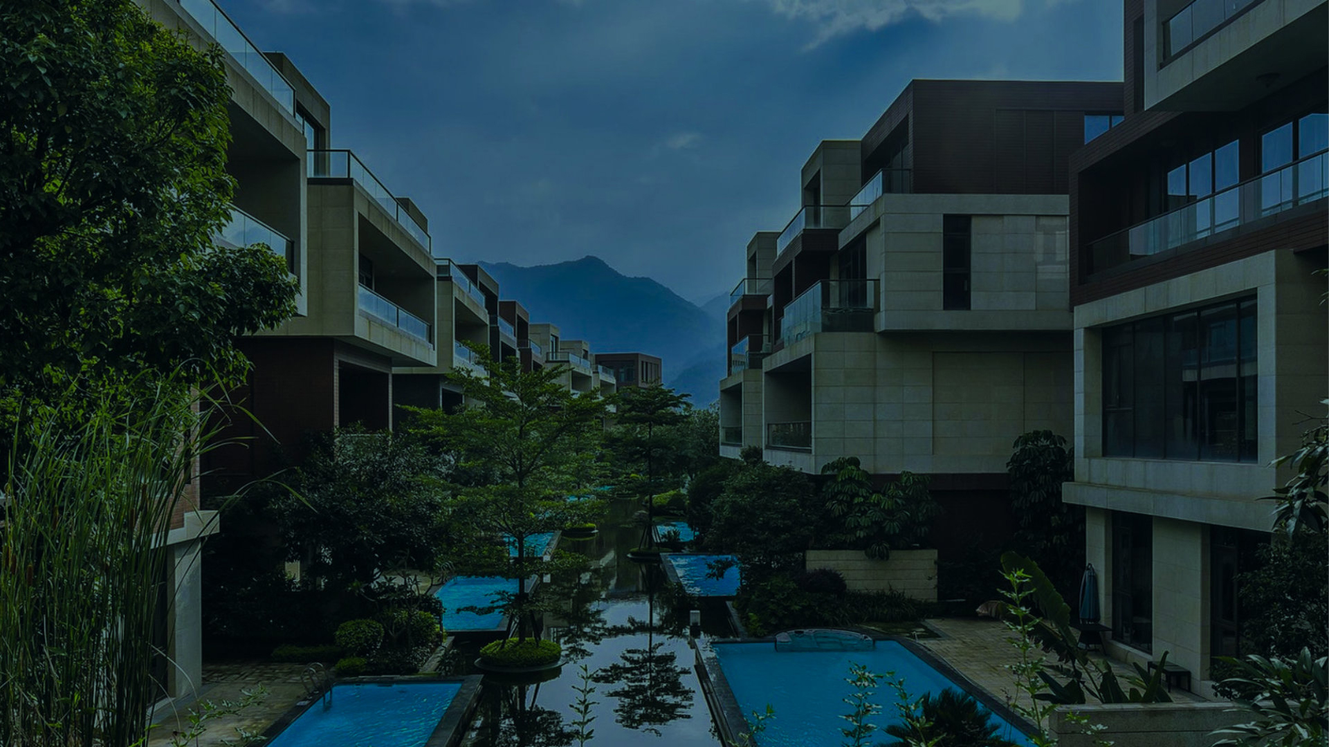 Modern multi-story residential buildings with balconies overlooking outdoor pools and greenery at dusk, with mountains in the distance