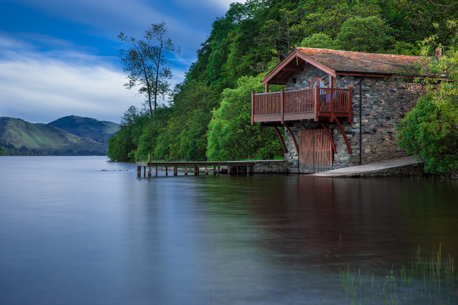 A stone boathouse with a wooden balcony on a calm lake shoreline surrounded by trees and hills