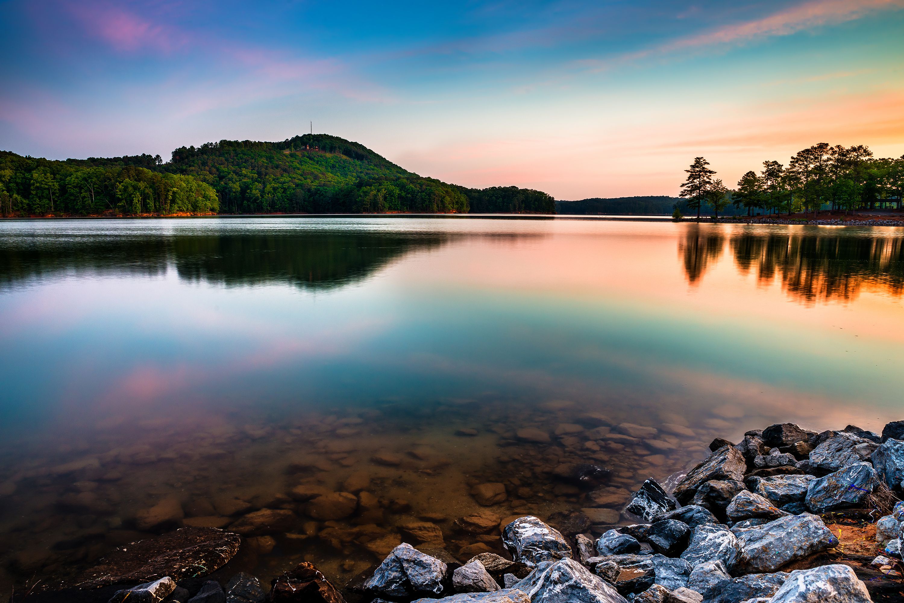 A calm lake at sunset with forested hills reflected on the water and rocks in the foreground